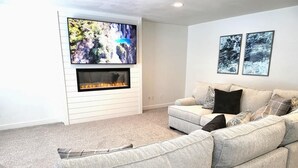 A living room with a fireplace, a large flat-screen TV, and a grey sofa. - Close to Marina & Market · 3BR House · Sleeps 10 (Garden City)