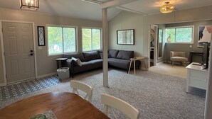 A spacious living room with a large window, a sofa, a dining table with chairs, and a small side table. - Cozy 2BD Retreat Near Danceland in Manitou Beach (Manitou Beach)