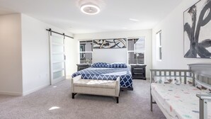 A bedroom with a large bed, a bench, a crib, and a wall-mounted shelf.