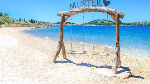 Un columpio de madera con la palabra “MURTER” y un símbolo de corazón, situado en una playa arenosa con agua azul cristalina y colinas al fondo.