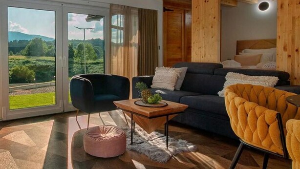 En moderne stue med sofa, stole, et sofabord og udsigt til et grønt landskab gennem store vinduer.