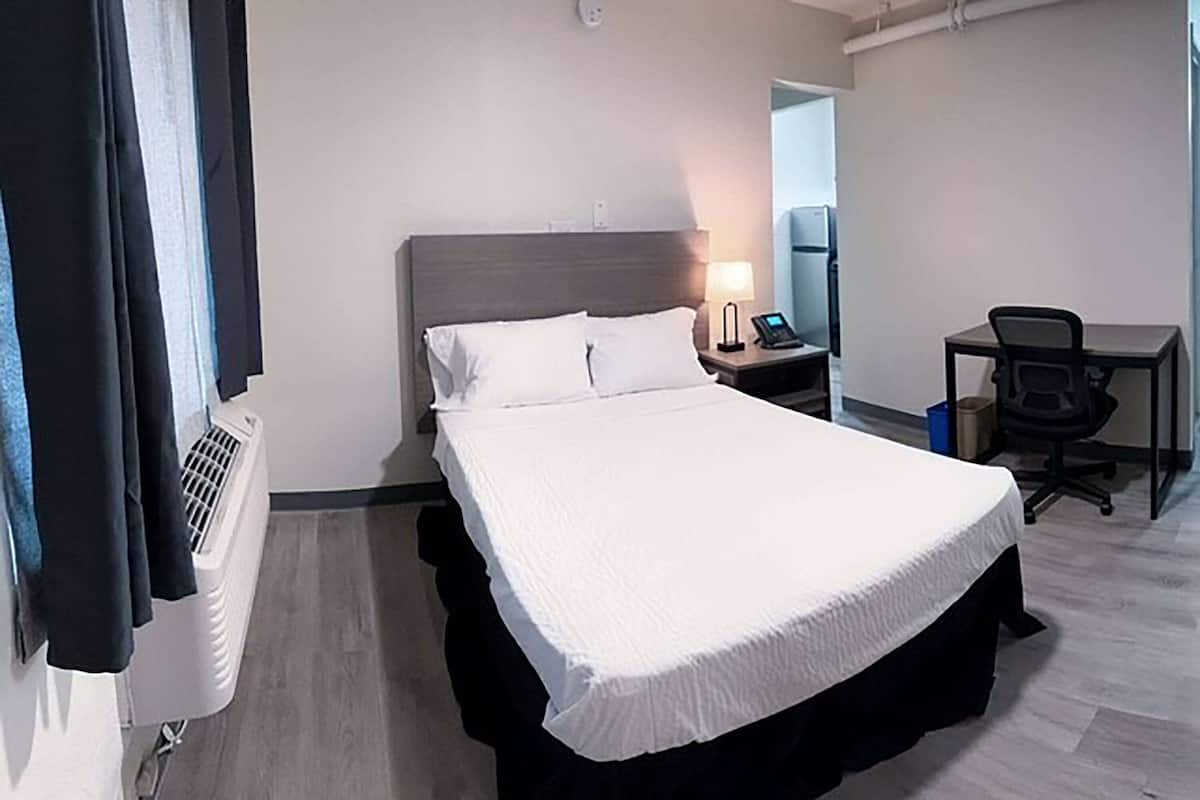 Quarto de hotel com cama, mesa de cabeceira, luminária, poltrona e escrivaninha com cadeira.