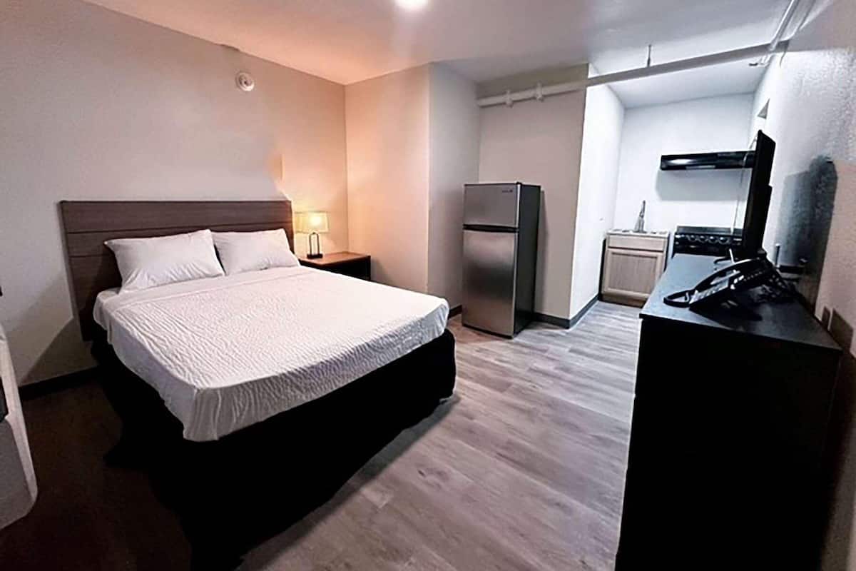 Quarto com cama, mesa de cabeceira, luminária, geladeira e uma área de cozinha compacta.