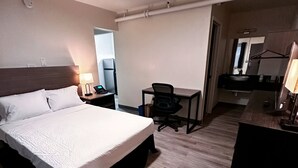 A hotel room with a bed, desk, chair, and a bathroom with a mirror and sink. - Studio City Suites, A Travelodge By Wyndham (Texarkana)