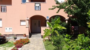 A building with a central entrance, a number '2' above the door, and a garden with flowers and plants. - Apartmani Luna - One Bedroom  Apartment (Sukosan)