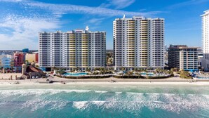 Un hotel al costat de la platja amb diversos edificis alts, una zona de piscines i la platja en primer pla.