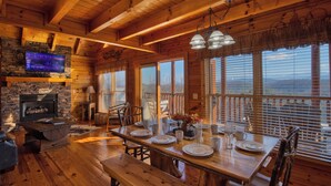 L’intérieur d’un chalet en bois est agrémenté d’une cheminée en pierre, d’une table à manger dressée pour quatre personnes et de grandes fenêtres équipées de stores.