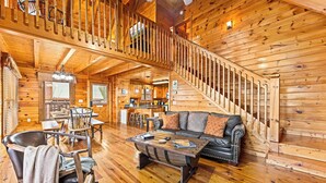 L’intérieur de cette vaste cabane en rondins offre un espace de vie central agrémenté d’un canapé, d’une table basse et d’une table à manger. Un escalier avec une rampe en bois mène à l’étage supérieur.