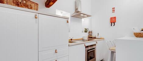 Cocina compacta con armarios blancos, horno integrado, campana extractora de acero inoxidable y una pequeña encimera con fregadero.