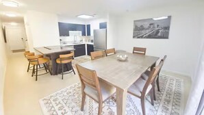 A dining area with a table and chairs, a kitchen in the background, and a cityscape painting on the wall.