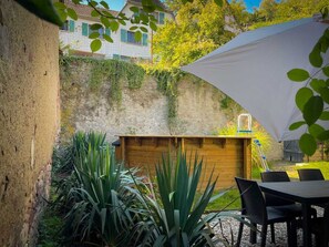 Un espace patio avec une table et des chaises en bois, un parasol blanc et un mur de pierre orné de plantes grimpantes.