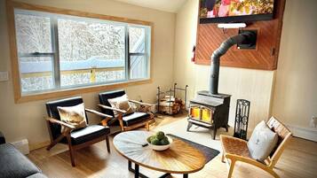 Una sala de estar acogedora con chimenea, asientos cómodos y un ventanal con vistas a árboles cubiertos de nieve.