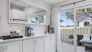 A modern kitchen with white cabinets, a coffee machine, and a window overlooking a patio area with a white fence and outdoor seating. - Hide Tide Hideaway at Carlin Park (Jupiter)