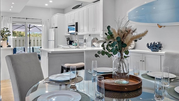 A modern kitchen with white cabinets, a dining area with a glass table set for four, and a view of the outdoors. - Hide Tide Hideaway at Carlin Park (Jupiter)