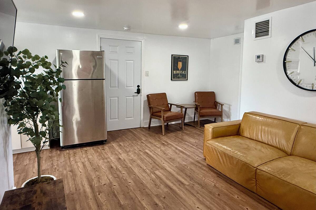 A compact living space with a wooden floor, a refrigerator, a sofa, two chairs, a small table, and a wall clock.