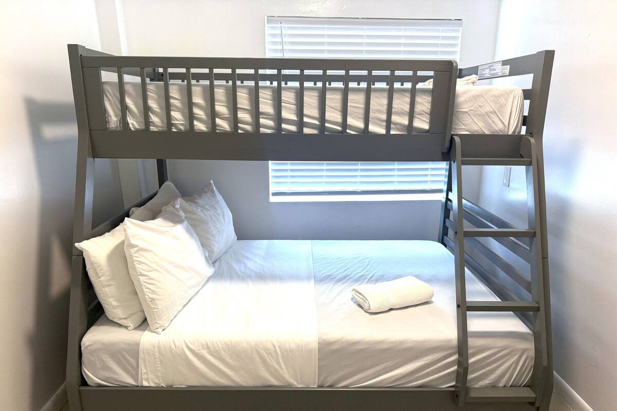 A bunk bed with white bedding and a ladder, a window with blinds, and a white wall.