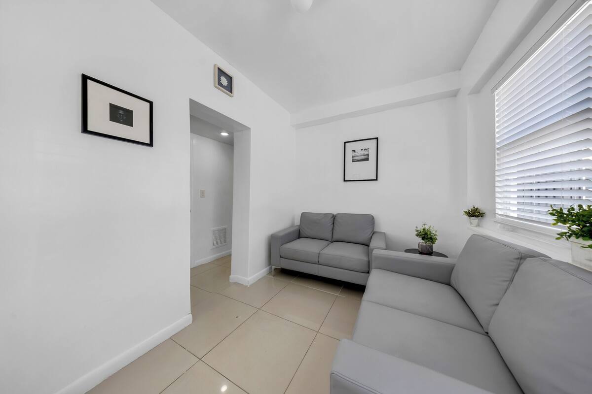 A modern living room with a grey sofa, a small table with a plant, and a window with blinds.