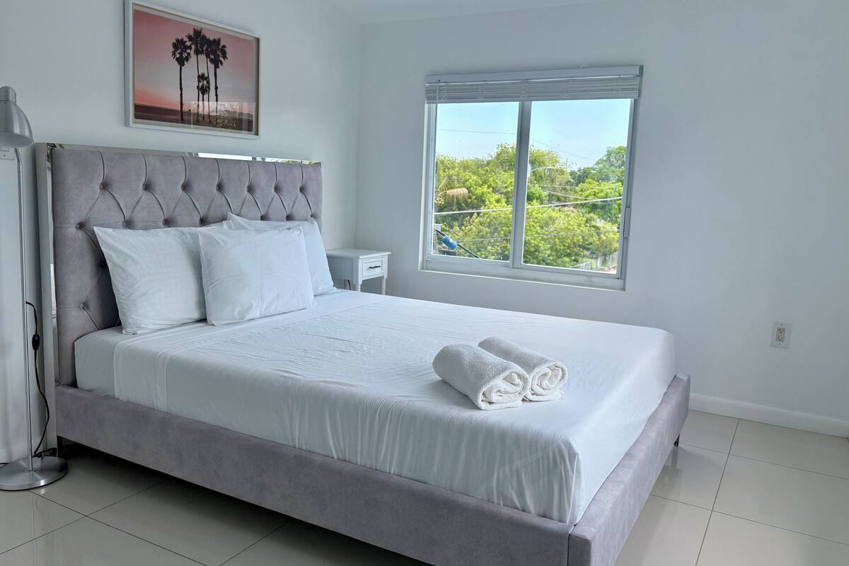 A neatly made bed with white linens and pillows, a grey upholstered headboard, a bedside table, a framed picture on the wall, and a window with a view of greenery.