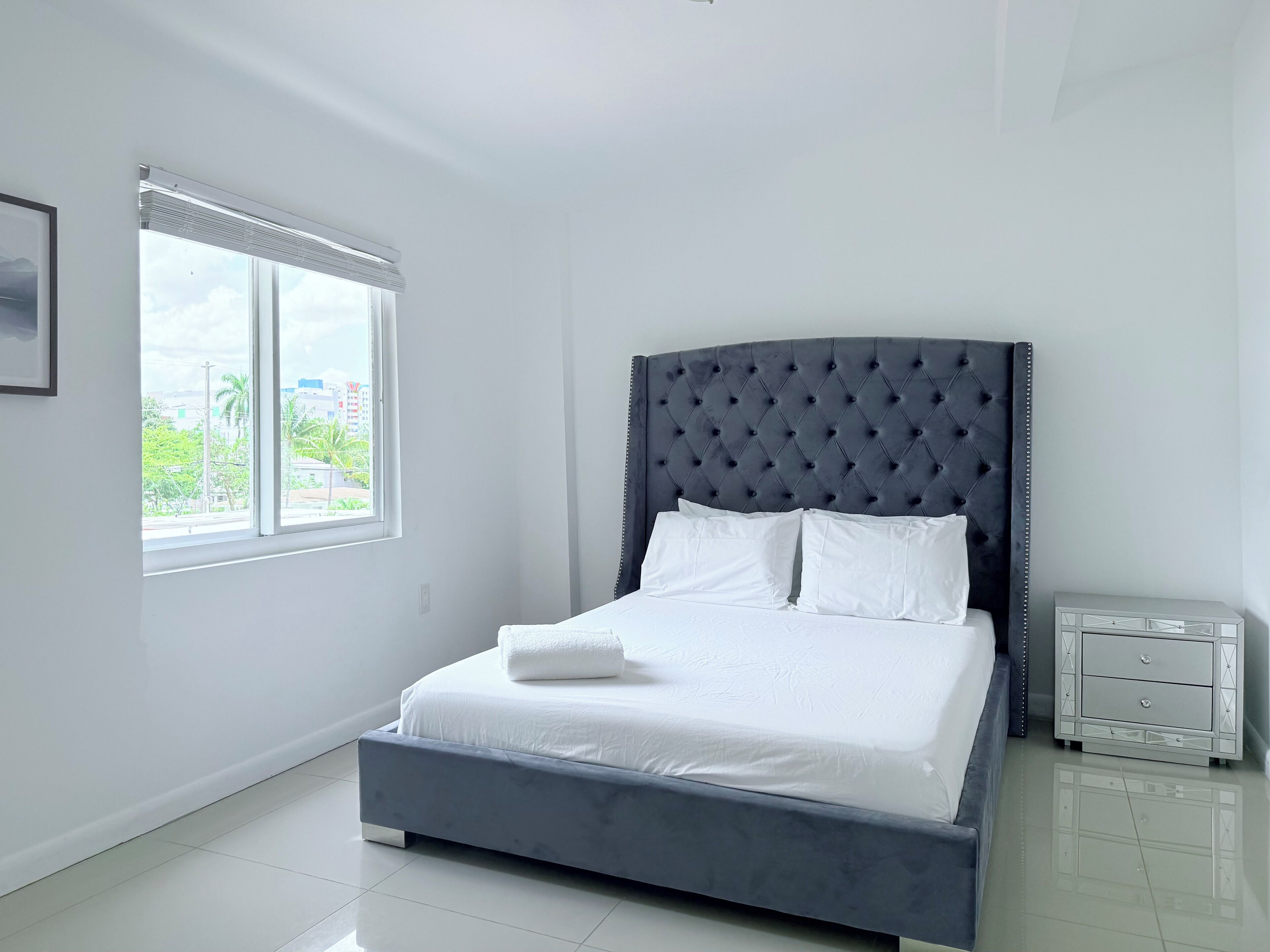 A bedroom with a large bed, a window with blinds, a bedside table, and a framed picture on the wall.