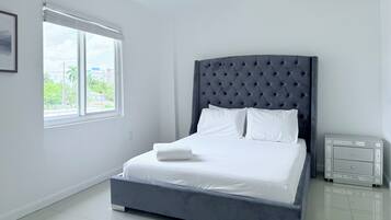 A bedroom with a large bed, a window with blinds, a bedside table, and a framed picture on the wall.