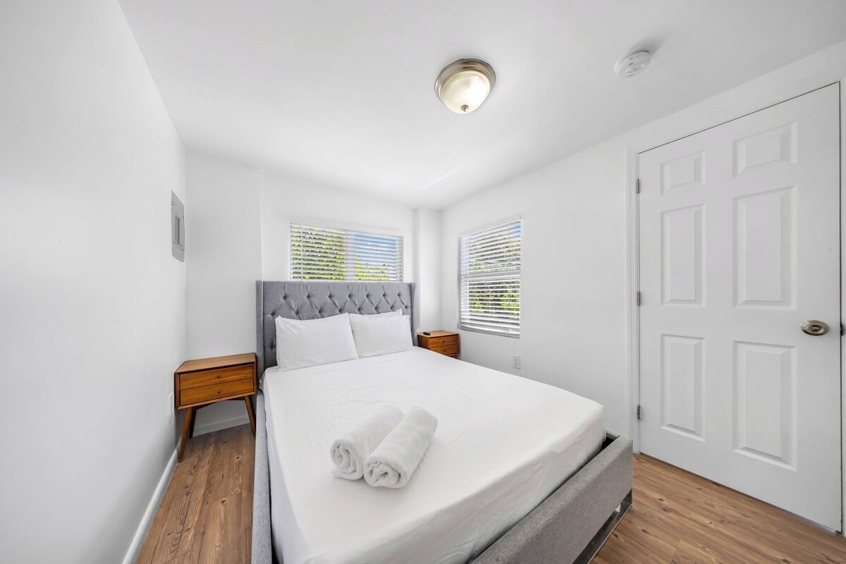 A neatly made bed with a grey headboard, white bedding, and rolled towels. There are two bedside tables with wooden finishes, a white door, and a window with blinds.