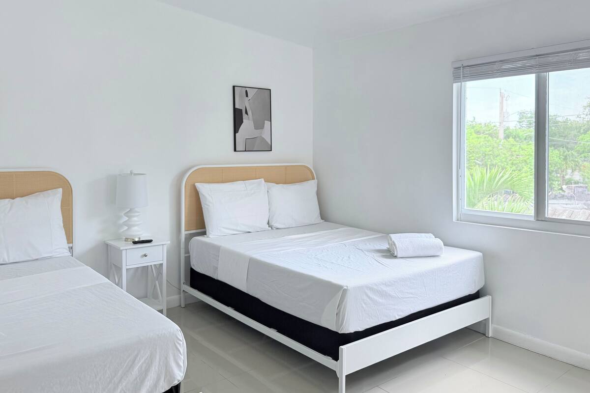 A hotel room with a bed, a bedside table, a lamp, and a window with a view of greenery.