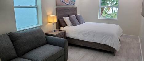 Un dormitorio con cama, una mesita de noche, una lámpara, un sofá gris y una ventana con vistas al exterior.