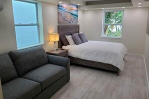 A bedroom with a bed, a nightstand, a lamp, a gray sofa, and a window with a view of the outdoors.