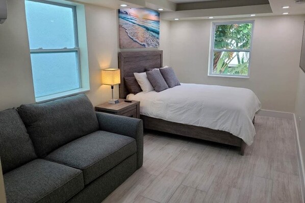 A bedroom with a bed, a nightstand, a lamp, a gray sofa, and a window with a view of the outdoors.