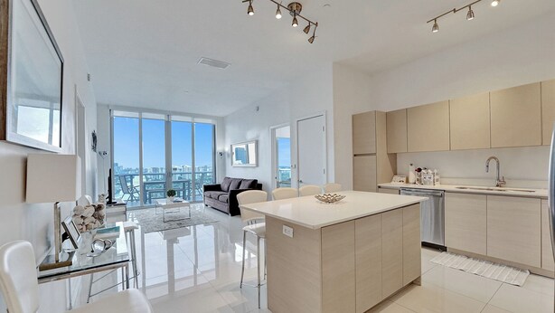 A modern kitchen with a central island, a dining area with a glass table, and a living room with a sofa and large windows.