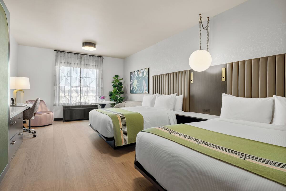 A hotel room with two beds, a desk, a chair, a plant, and a window with curtains.