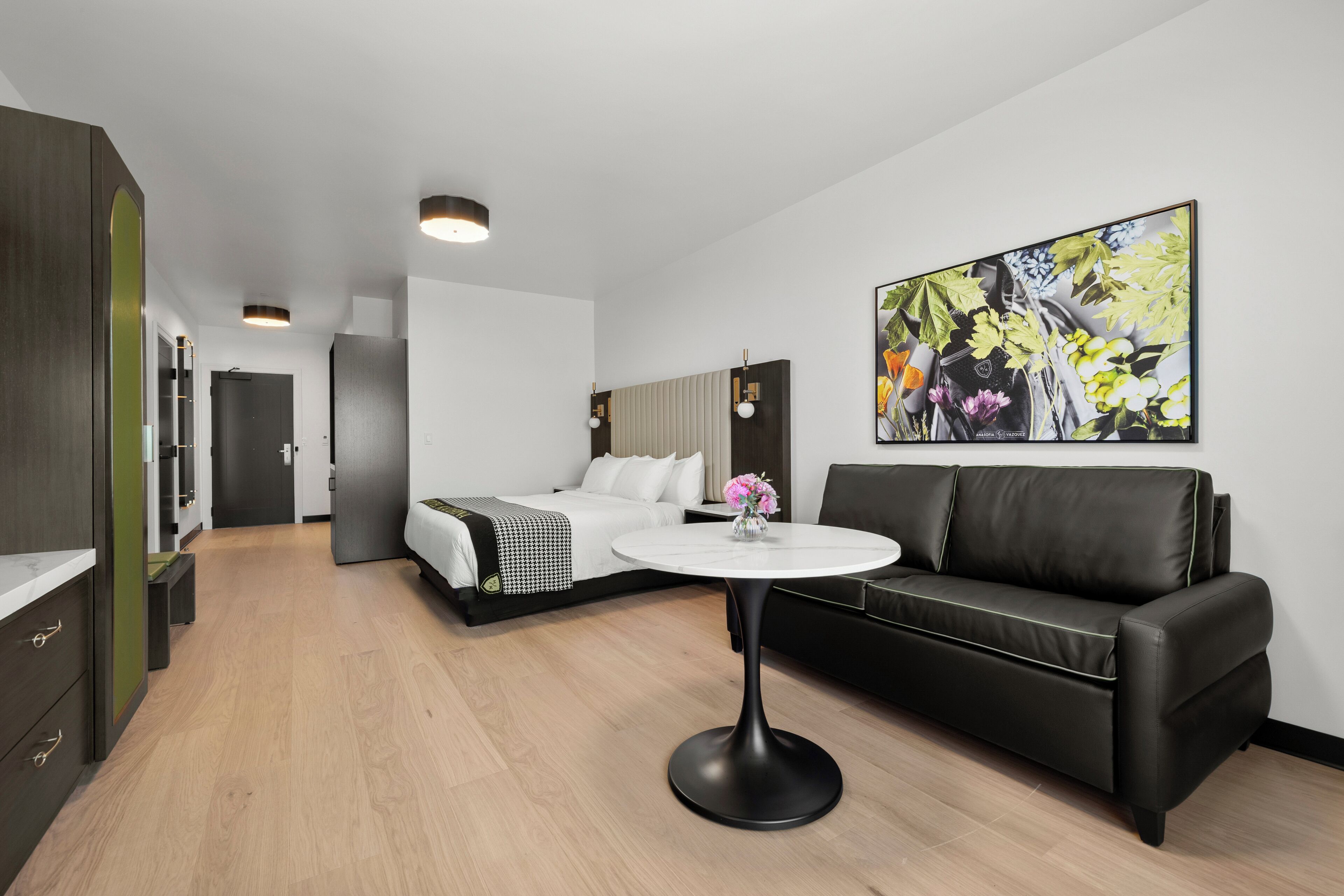 Une chambre d’hôtel moderne avec un lit, un canapé en cuir noir, une table ronde et une grande œuvre d’art florale accrochée au mur.