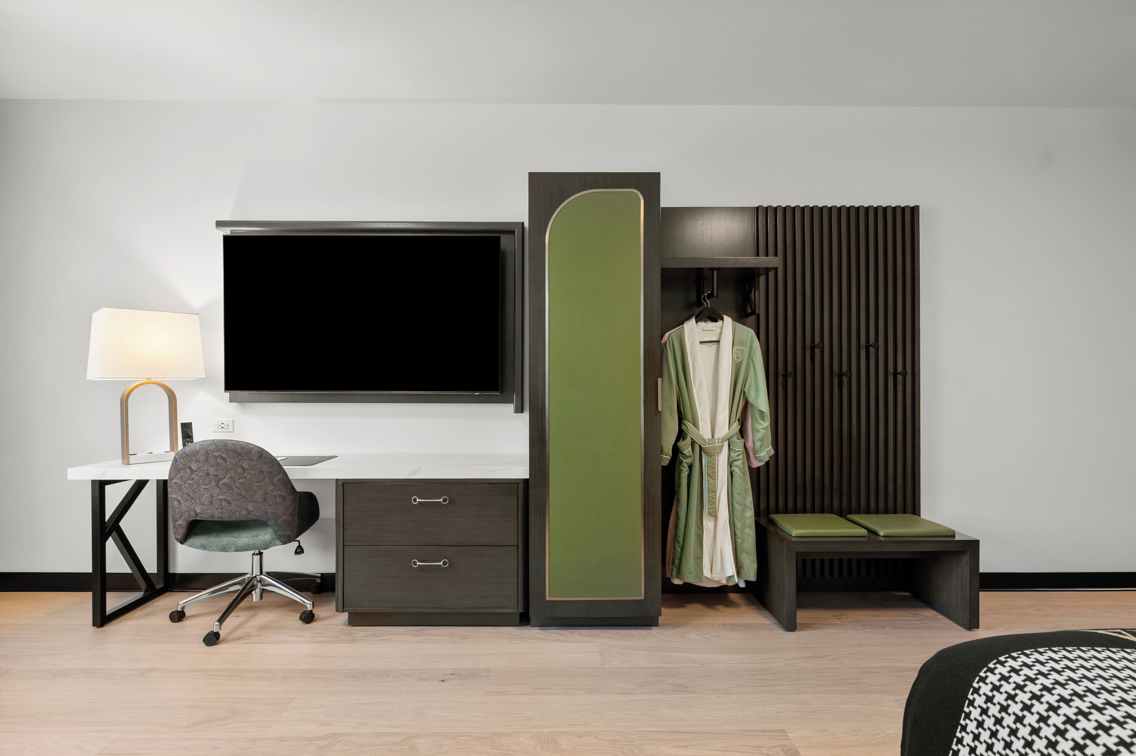 Ein modernes Hotelzimmer mit einem Flachbildfernseher, einem Schreibtisch mit Lampe, einem grauen Bürostuhl, einem grünen Bademantel und einer Bank mit grünen Kissen.