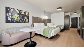 Quarto de hotel moderno com sofá, mesa redonda, cama com manta verde e um grande quadro floral na parede.