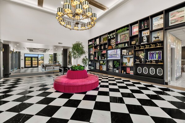 A hotel lobby with a black and white checkered floor, a central pink seating area, and a large chandelier.