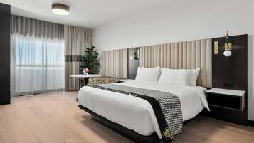 Una habitación de hotel moderna con una cama grande, mesitas de noche, una pequeña mesa auxiliar, una planta en maceta y un ventanal con cortinas.