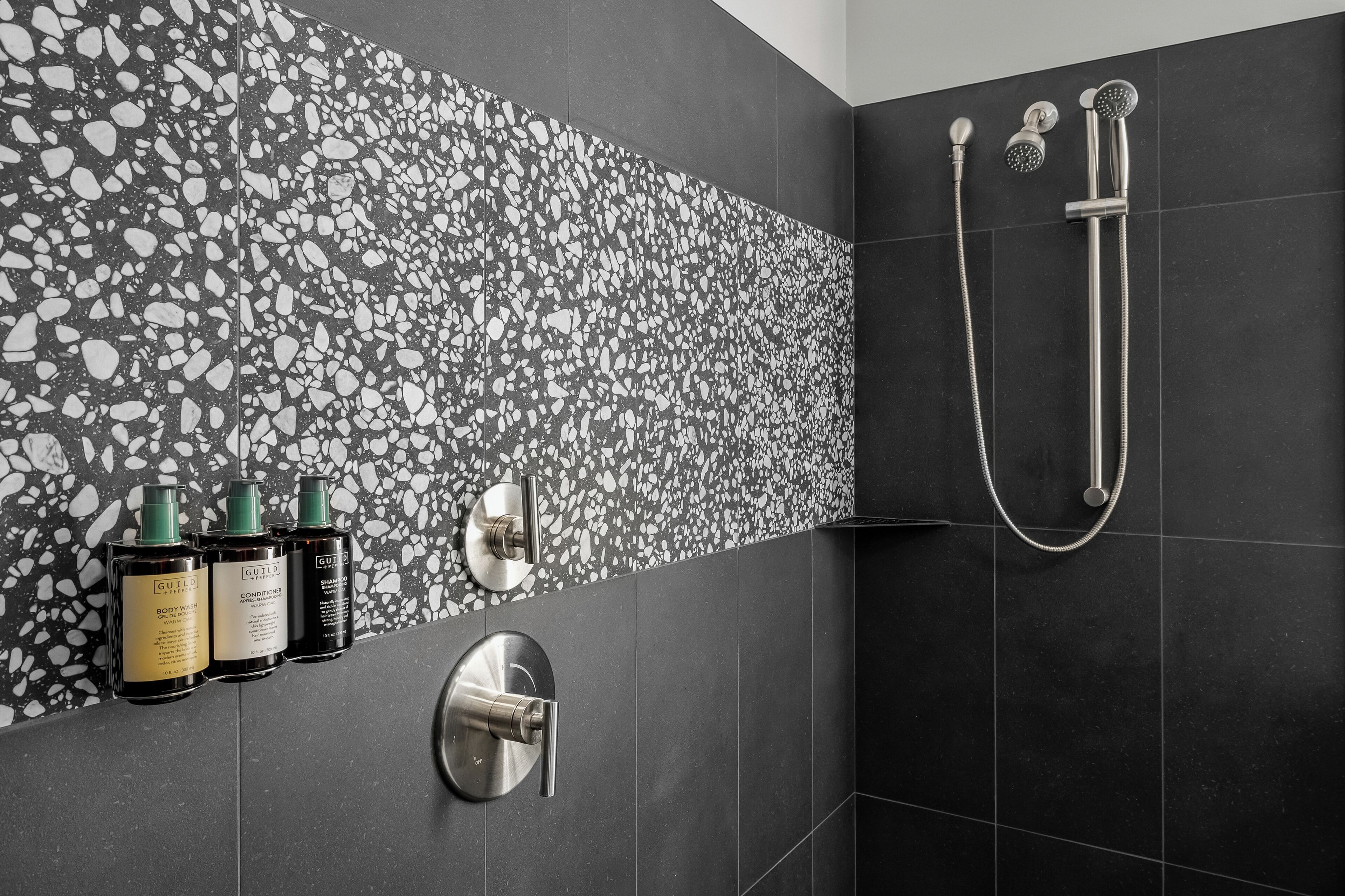 Une salle de bain moderne avec un mur décoré d’un motif de galets, une douchette à main et deux bouteilles de shampoing et d’après-shampoing.