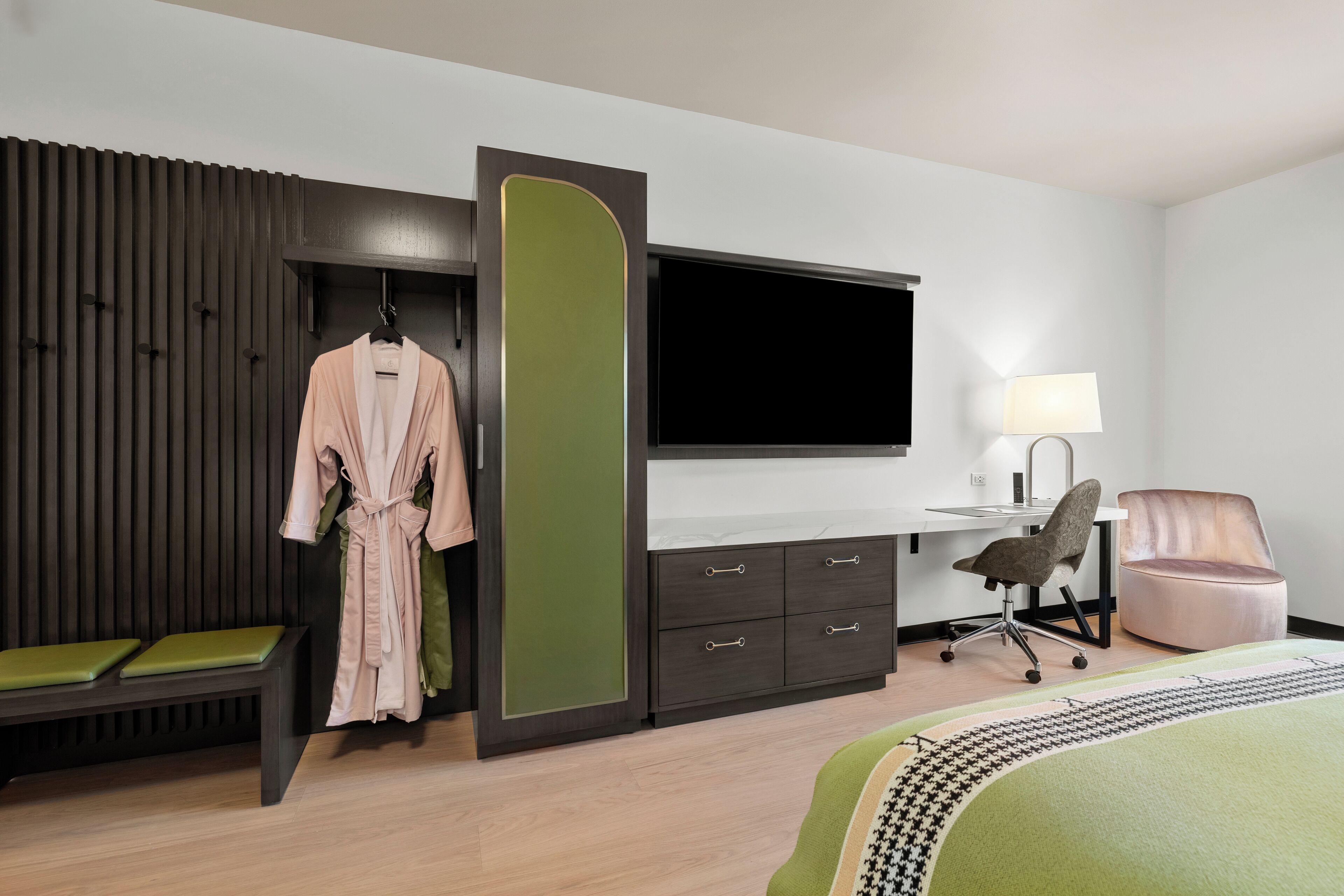 Una habitación de hotel moderna con una cama, un escritorio con una silla, un televisor, un banco y una bata colgada en un perchero.