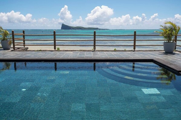 Ein klarblaues Schwimmbecken mit Holzterrasse und Geländer, mit Blick auf einen Strand und den Ozean mit einer entfernten Insel.