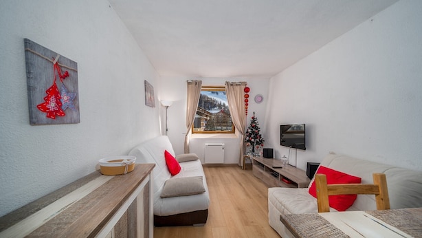 A cozy living room with a sofa, a wooden table, a TV, and a window with curtains. - Studio in Allos with Stunning Mountain Views (Allos)