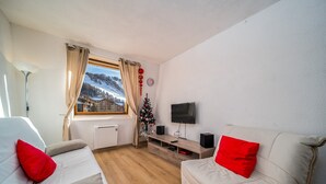 A cozy room with two white sofas, each with a red pillow, a small Christmas tree, a flat-screen TV on a wooden stand, and a window with curtains. - Studio in Allos with Stunning Mountain Views (Allos)
