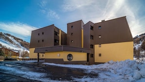 A modern building with a yellow facade and brown roof, featuring multiple windows and a circular window on the ground floor. - Studio in Allos with Stunning Mountain Views (Allos)