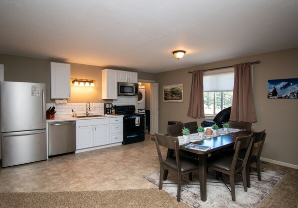 A compact kitchen with white cabinets, a microwave, a dishwasher, and a refrigerator. There is a dining table with chairs and a rug in the center.