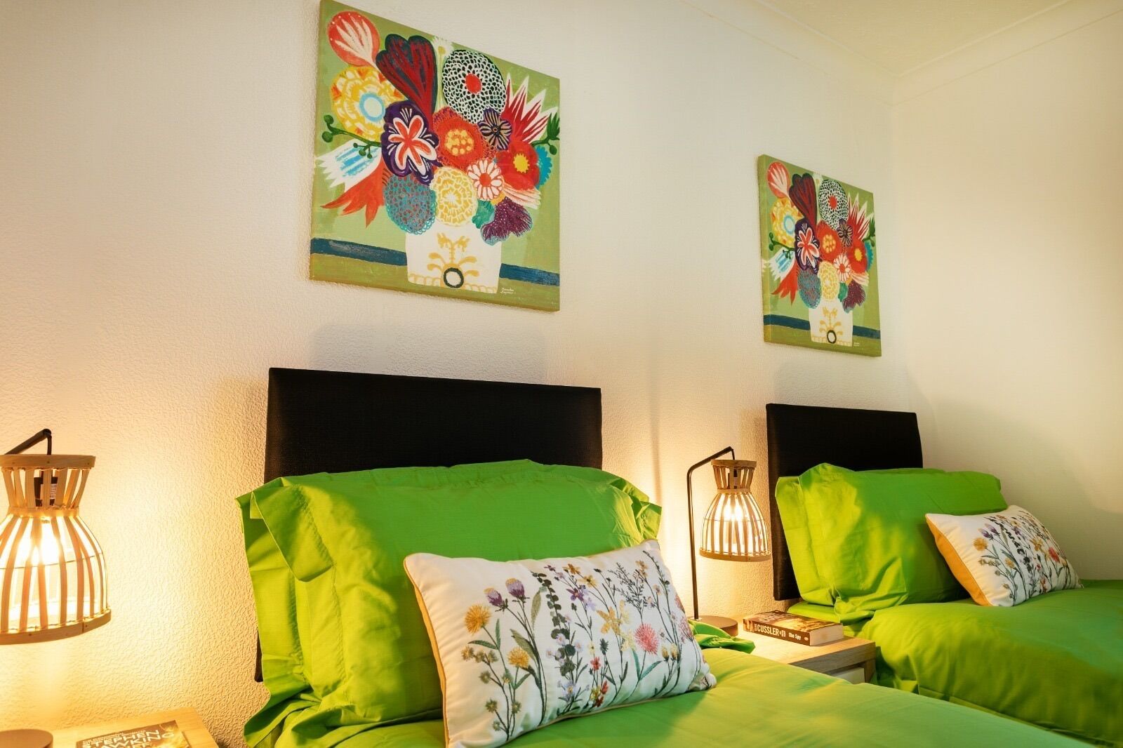 A room with two beds, each with green bedding and floral pillows. There are two wall-mounted paintings above the beds, and two table lamps on either side of the beds.