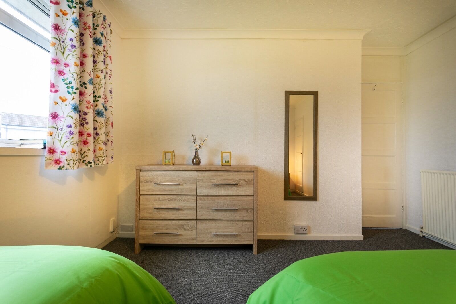 A bedroom with a wooden dresser, two green beds, a mirror, and a window with floral curtains.