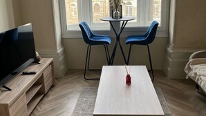 A living room with a TV on a wooden stand, a coffee table, two blue chairs, and a view of buildings through large windows. - Apartment - Parking - Pet Friendly - City Centre (Leeds)