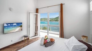 A hotel room with a bed, a TV on the wall, a view of the sea, and a balcony with curtains. - Stunning Pool Villa – Treasure of the Bay (La Trinité)