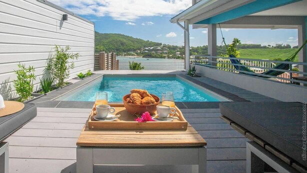 A poolside area with a wooden deck, a breakfast tray with croissants, and a view of a body of water and hills in the background. - Stunning Pool Villa – Treasure of the Bay (La Trinité)