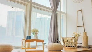 Una habitación con un ventanal grande, una silla de madera, un pouf tejido, una cesta tejida y un florero con flores.