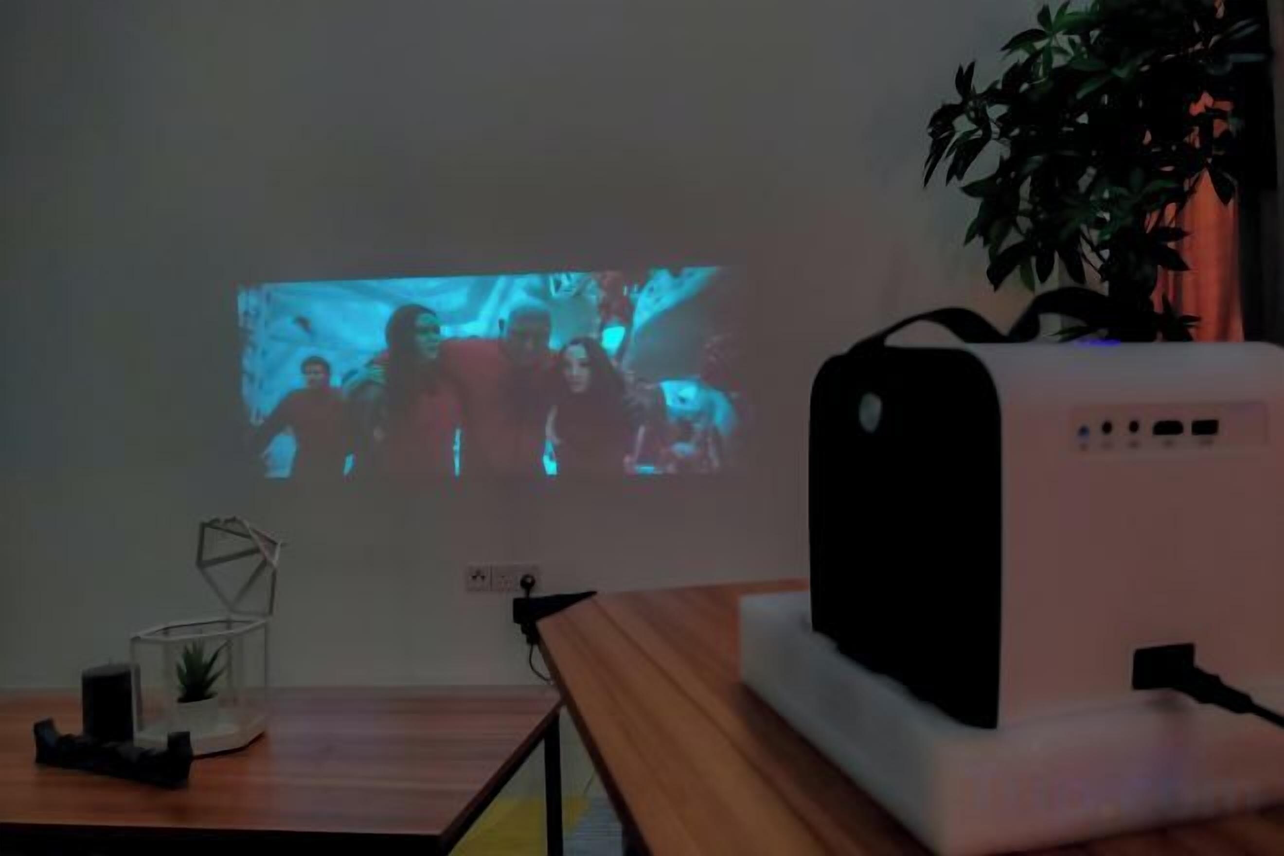 Un projecteur projette sur un mur l’image d’un groupe de personnes, un appareil blanc est posé sur une table, et une plante se trouve à l’arrière-plan.